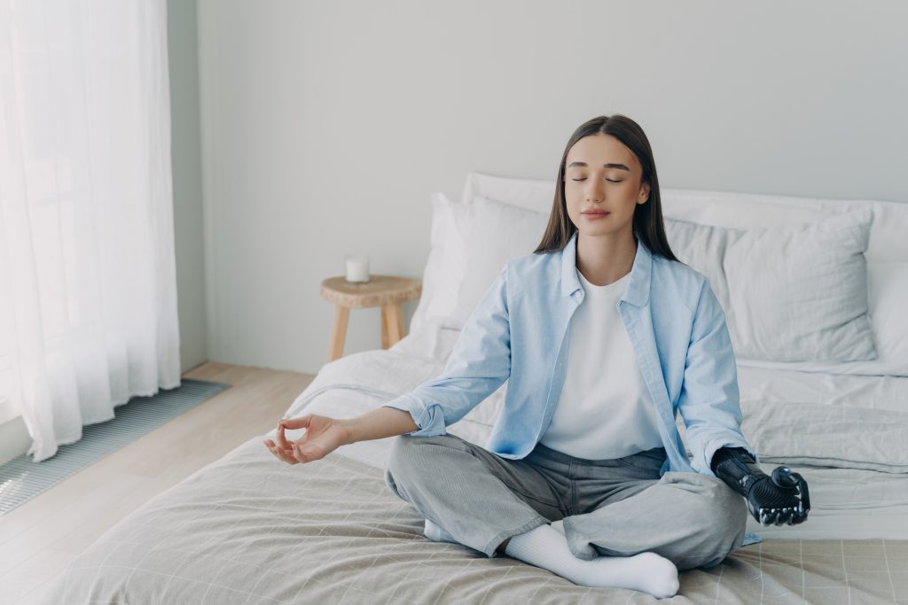 Frau sitzt mit geschlossenen Augen im Schneidersitz auf ihrem Bett und meditiert
