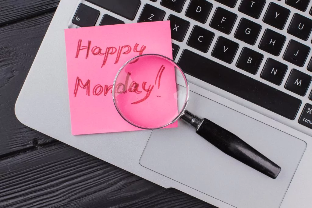 Laptop auf der eine Lupe liegt, die auf eine pinken Zettel mit der Aufschrift "Happy Monday" zeigt. Dunkler Holzhintergrund.