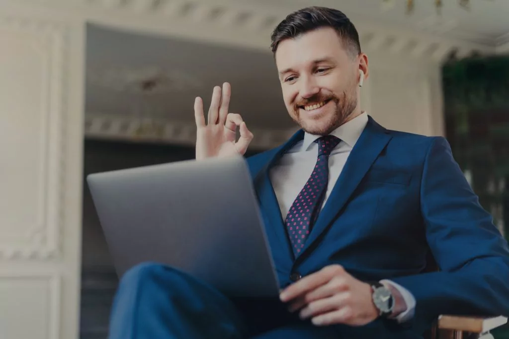 Geschäftsmann in blauem Anzug sitzt vor einem Tablet, schließt den Daumen mit dem Zeigefinger zusammen und lächelt dabei