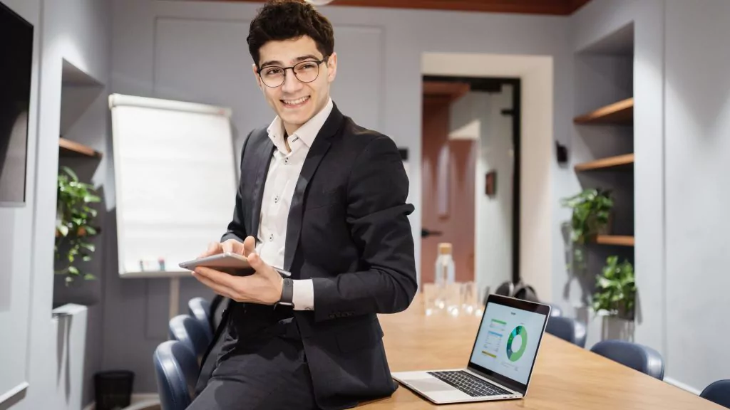 Mann mit Brille und Business-Outfit hält Taschenrechner in der Hand, lehnt sich an Tisch und lächelt