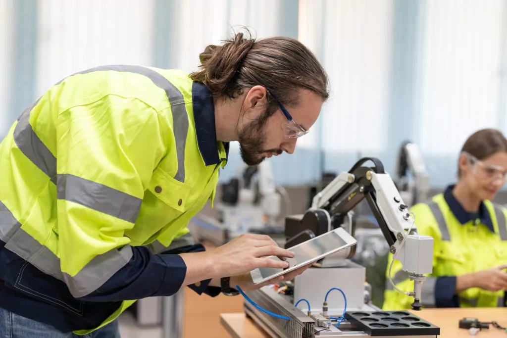 Mann in Arbeitsschutzkleidung hält ein Tablet in der Hand und beugt sich über einen Roboterarm