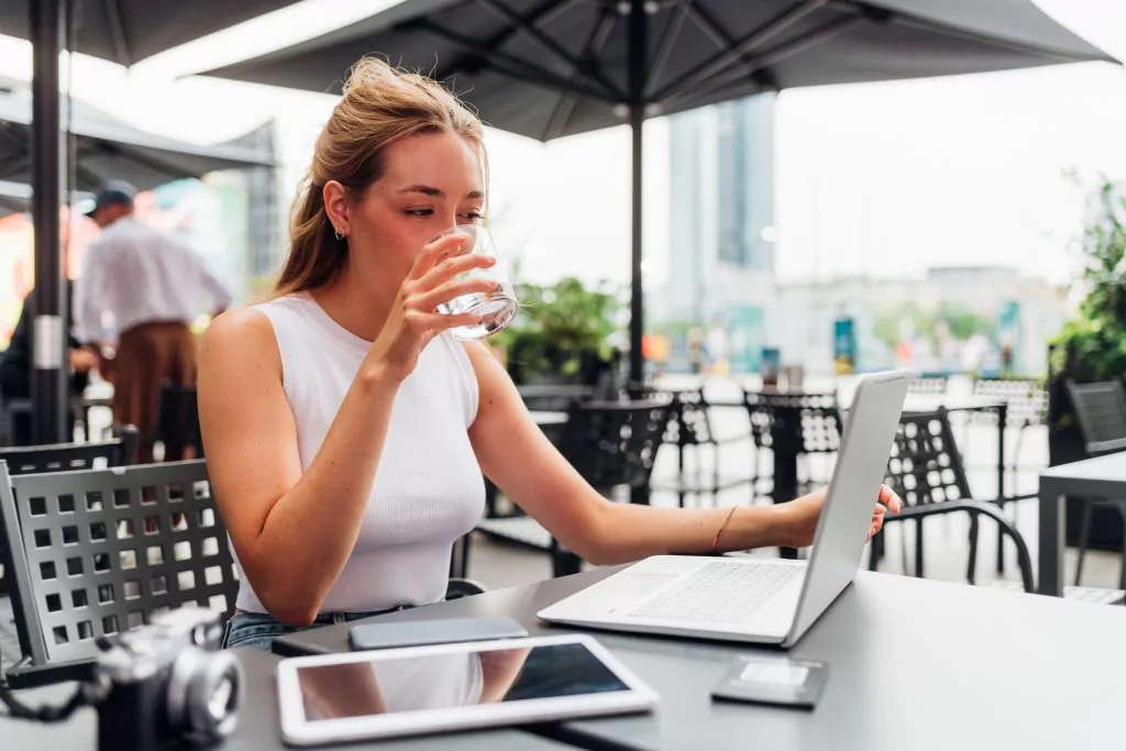 Junge Frau mit weißem Top hält ein Glas mit Wasser zu ihrem Mund. Sie sitzt in einem Kaffeehaus und hat ein Notebook vor sich aufgestellt