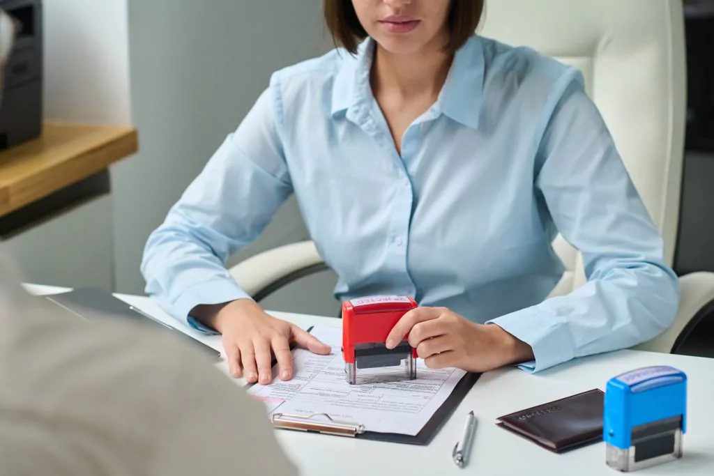Frau mit blauer Bluse sitzt an einem Schreibtisch und drückt einen roten Stempel auf eine Unterlage für eine Absage einer Bewerbung