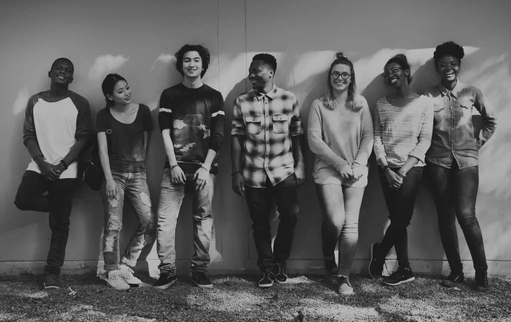Schwarz-weiß Aufnahme von sieben jungen Menschen unterschiedlicher Nationen, die an einer Wand gelehnt stehen