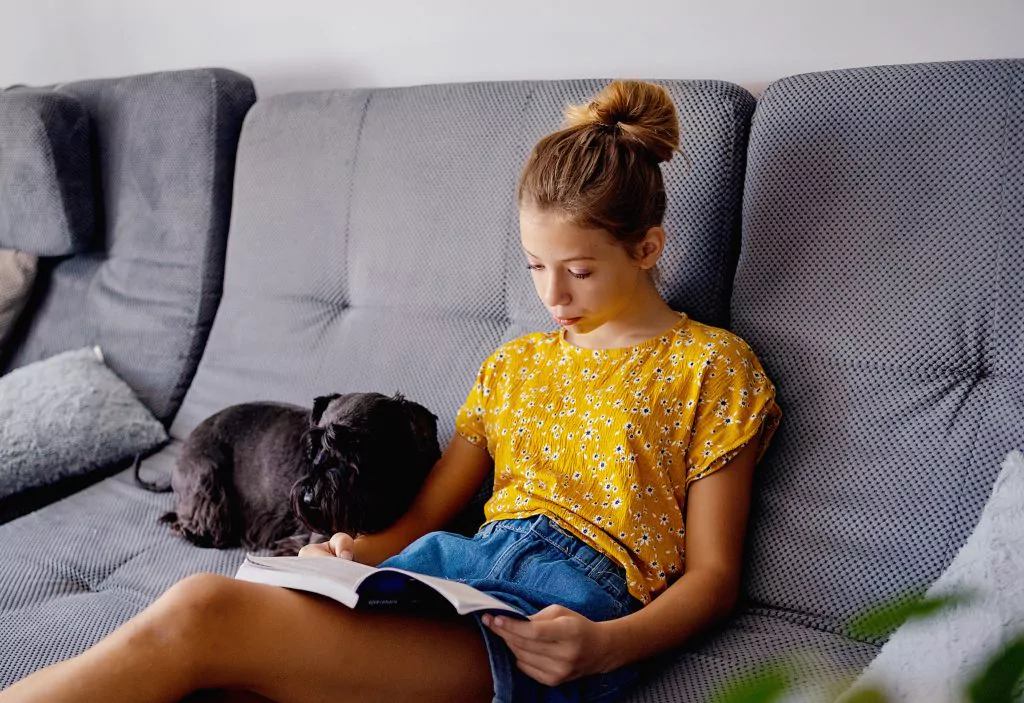 Junge Frau sitzt mit einem Buch und einem Hund auf einem Sofa