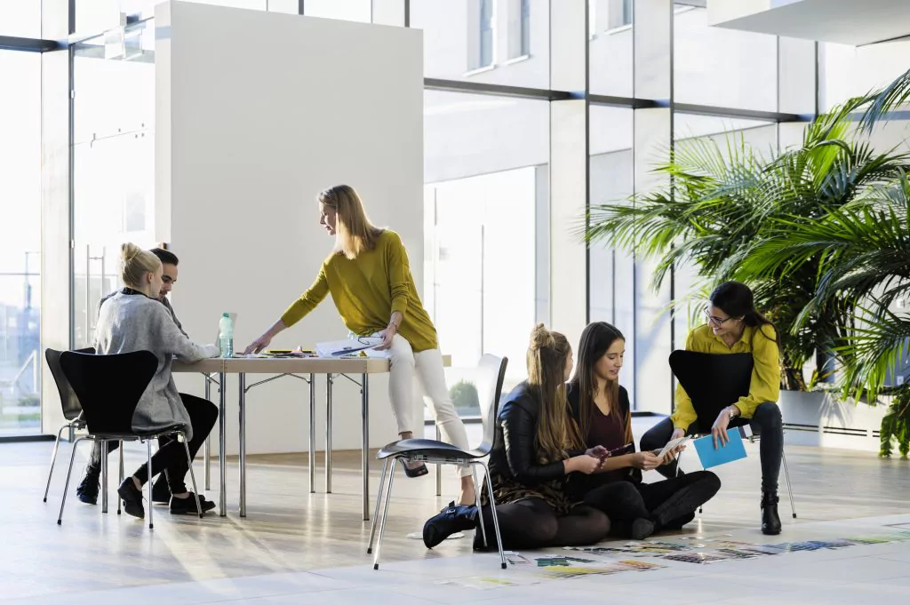 Zwei Menschen sitzen an einem Tisch. Eine Frau sitzt auf dem Tisch. Vor dem Tisch sitzen zwei junge Frauen auf dem Boden. Daneben eine Frau auf einem Sessel 