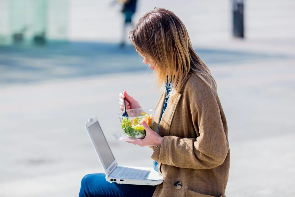 Frau sitzt mit Laptop im Freien und isst einen Salat